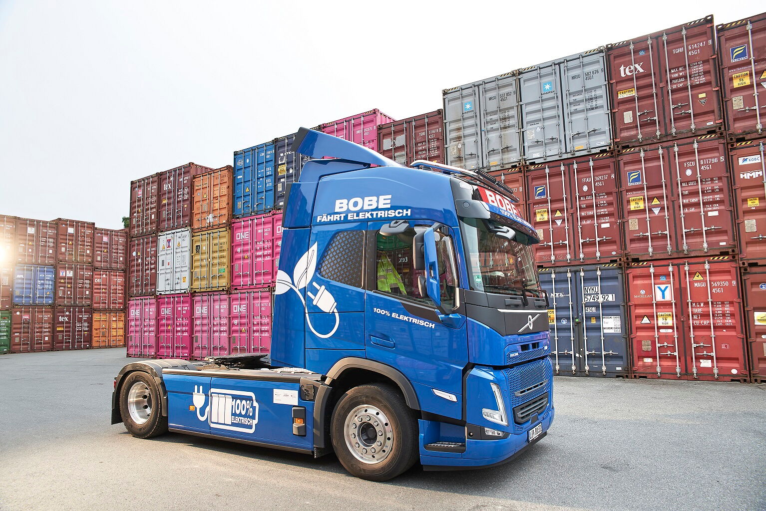 Ein blauer BOBE Elektro-LKW (Volvo FH Electric) vor einer imposanten Kulisse aus gestapelten Überseecontainern – BOBE