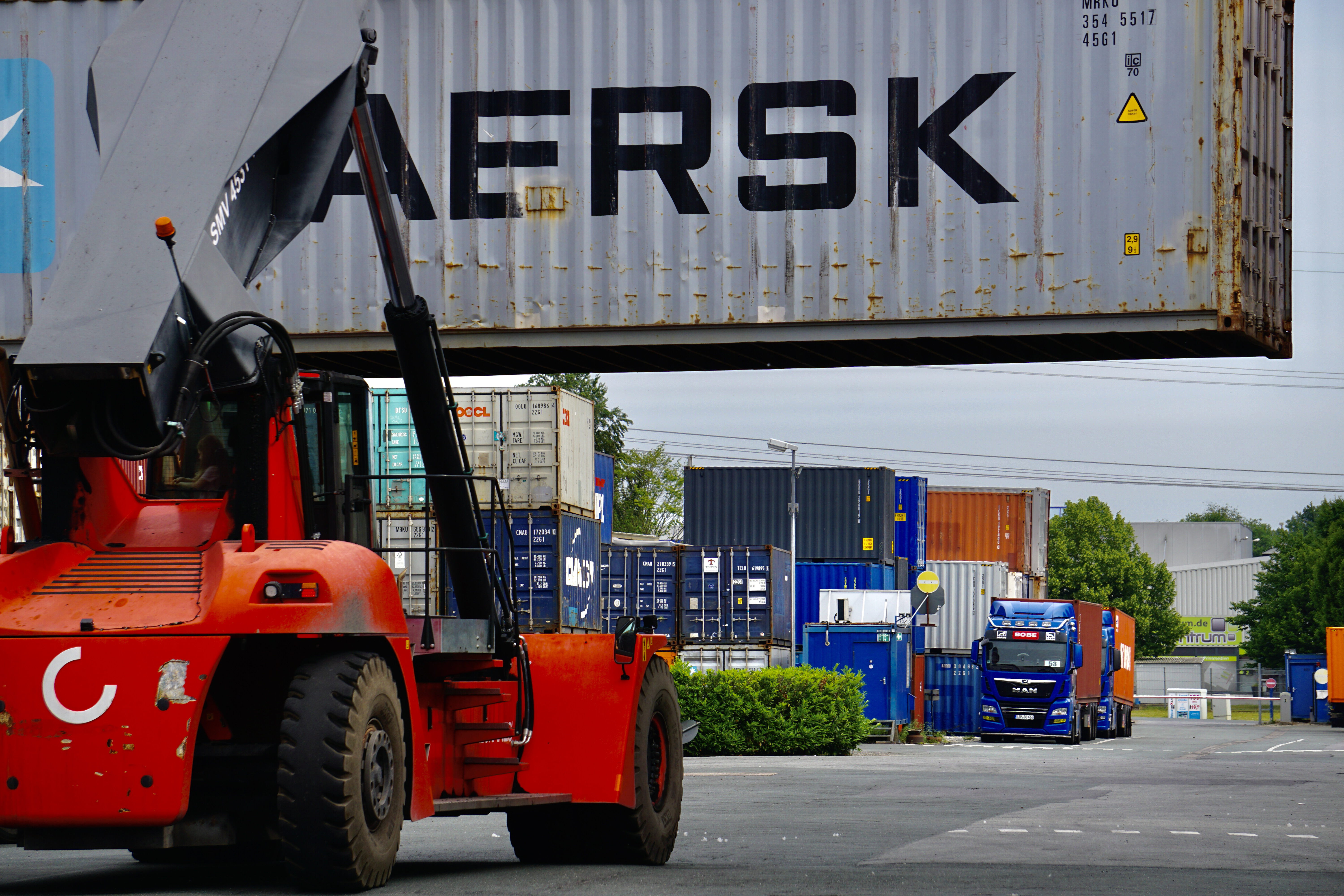 Ein Reachstacker hebt einen Maersk-Container; im Hintergrund wartet ein blauer BOBE LKW im Depot