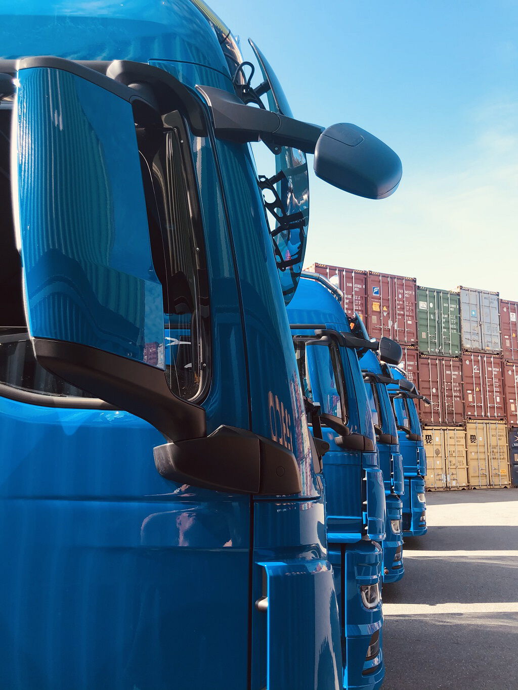 Perspektivische Ansicht einer Reihe blauer BOBE-LKWs vor einer Wand aus gestapelten Seecontainern auf dem Terminal – BOBE