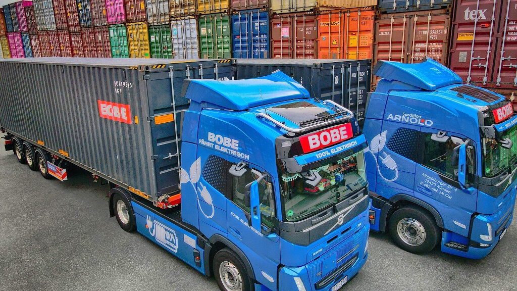 Zwei blaue Elektro-LKW der Speditionen BOBE und Arnold stehen mit einem beladenen Auflieger vor einer Wand aus bunten Seecontainern – BOBE