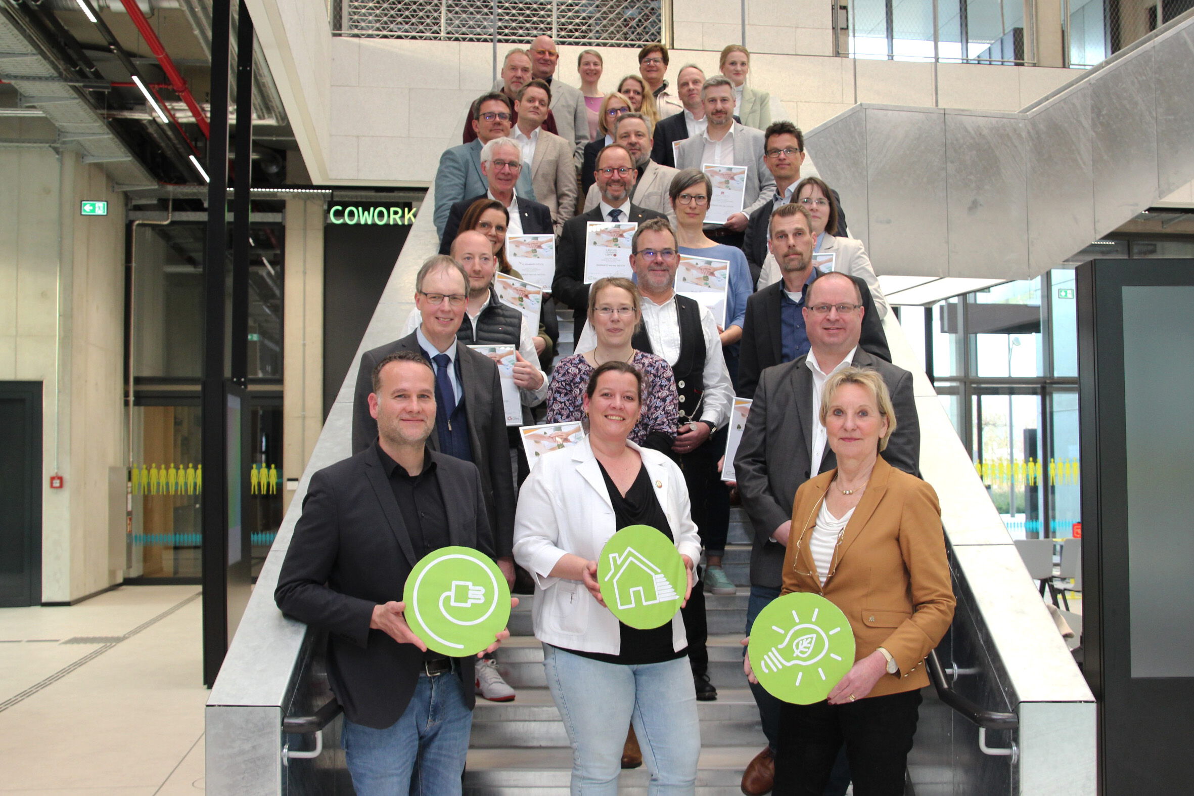Eine Gruppe von Projektbeteiligten posiert auf einer Treppe mit grünen Nachhaltigkeitssymbolen und Urkunden – BOBE