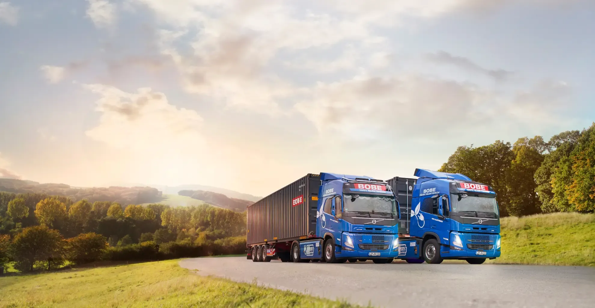 Zwei blaue LKW mit dunklen Containern stehen auf einer Straße in ländlicher Umgebung – BOBE