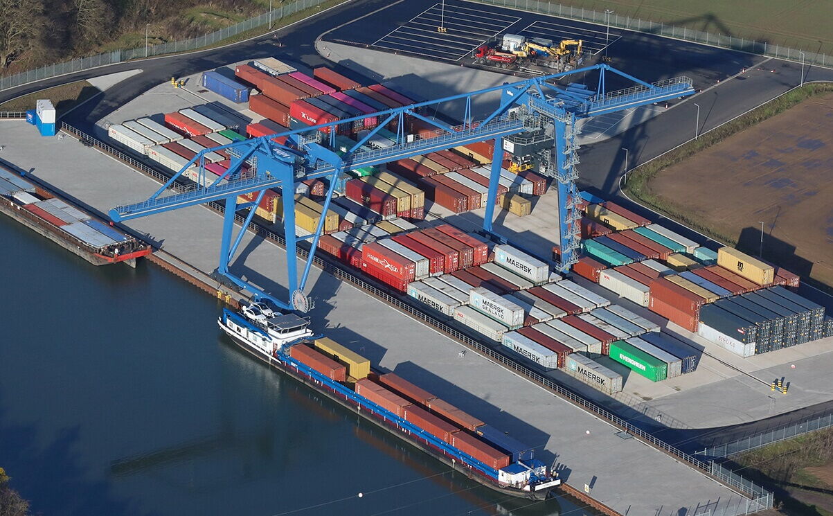 Luftaufnahme eines Containerterminals am Kanal mit einem großen blauen Portalkran bei der Beladung eines Binnenschiffs – BOBE