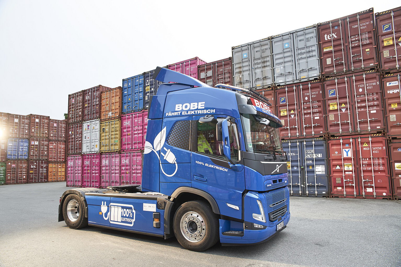 Ein blauer BOBE Elektro-LKW (Volvo FH Electric) vor einer imposanten Kulisse aus gestapelten Überseecontainern – BOBE