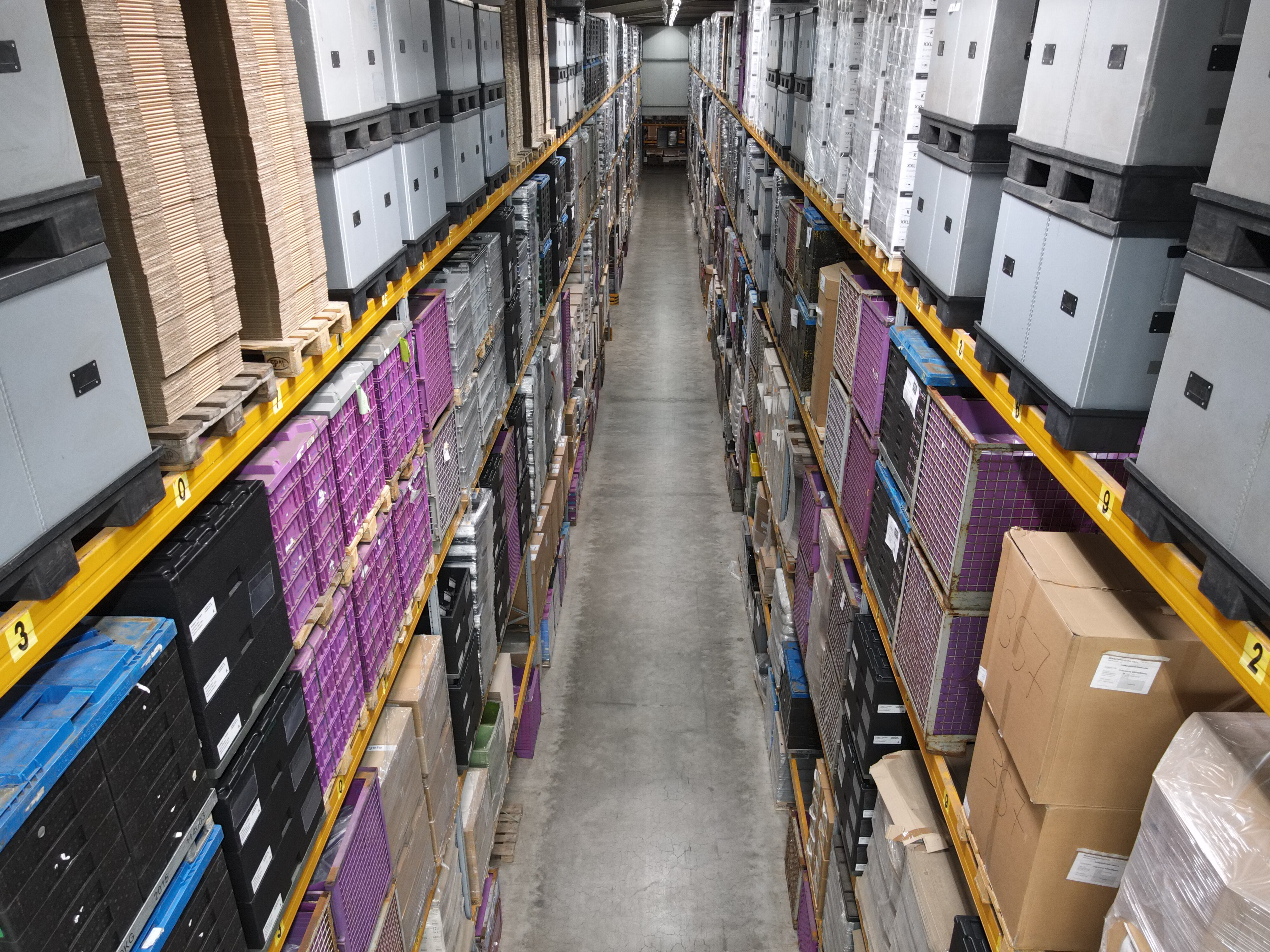 Blick von oben in einen schmalen Gang zwischen hohen Regalen mit diversen Transportboxen und Kartons – BOBE