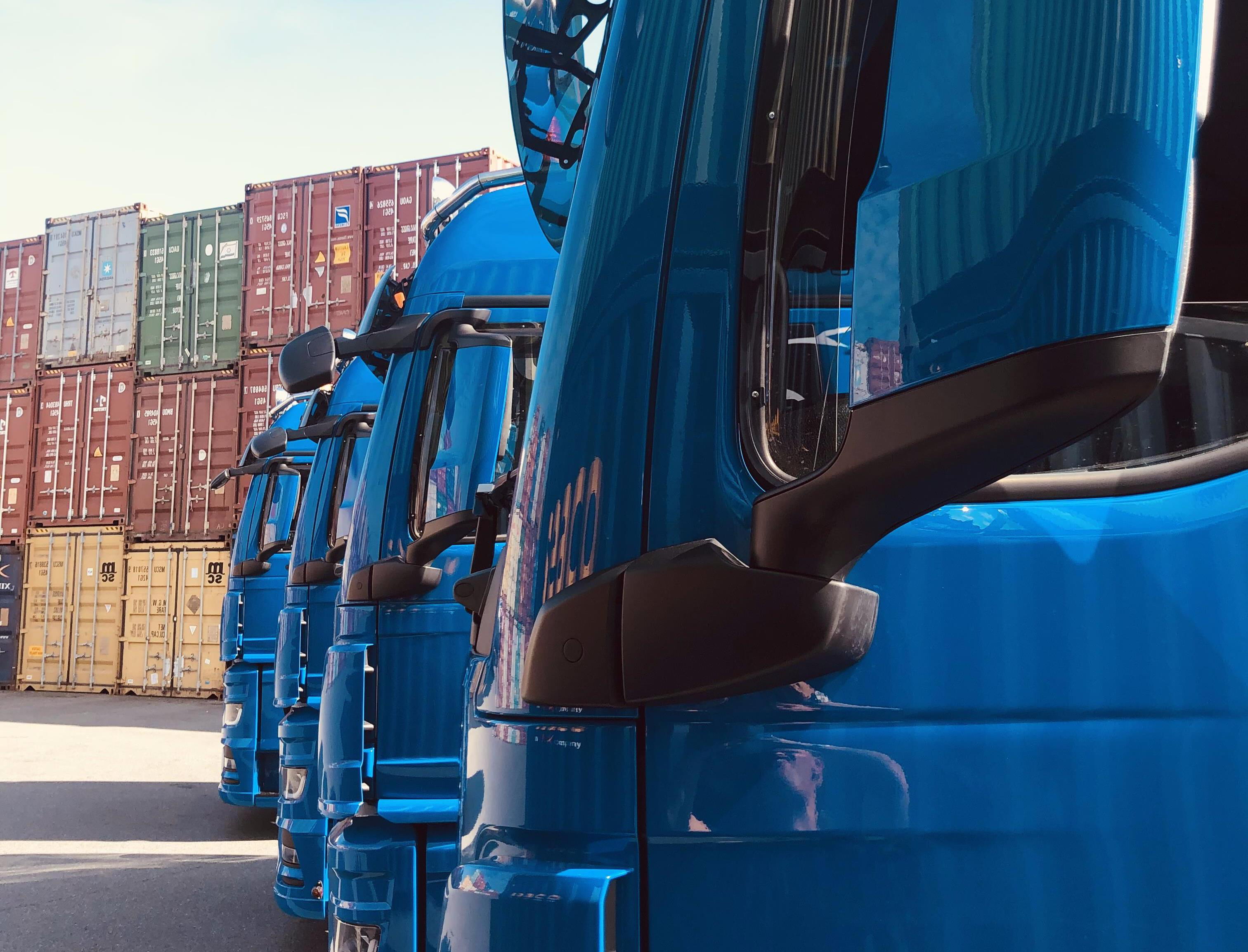 Detailaufnahme einer Reihe blauer BOBE-LKW vor einer Wand aus gestapelten Überseecontainern – BOBE