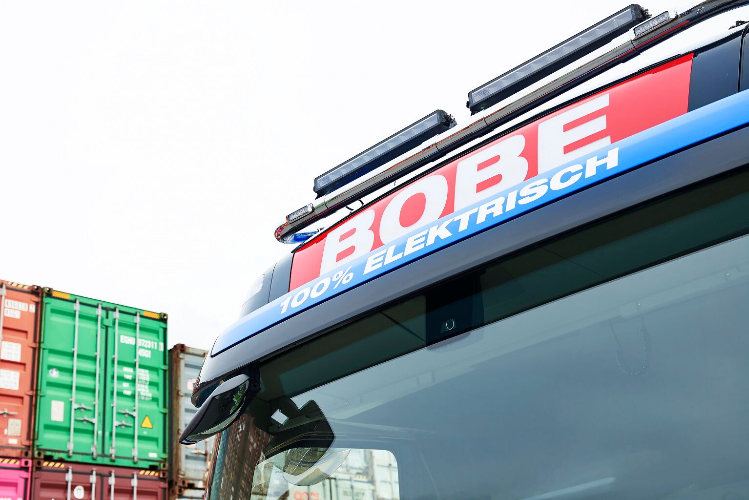 Detailansicht der Dachpartie eines LKWs mit dem Schriftzug „100% ELEKTRISCH“ vor gestapelten Containern – BOBE