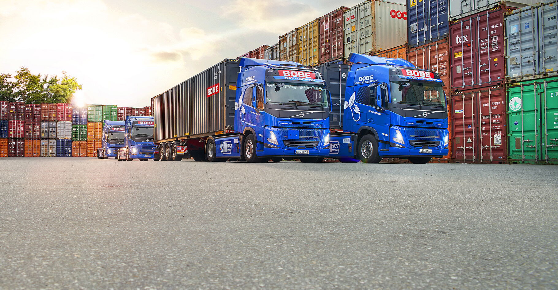 Blaue Elektro-LKW mit Trailern stehen vor einer Wand aus gestapelten Containern – BOBE