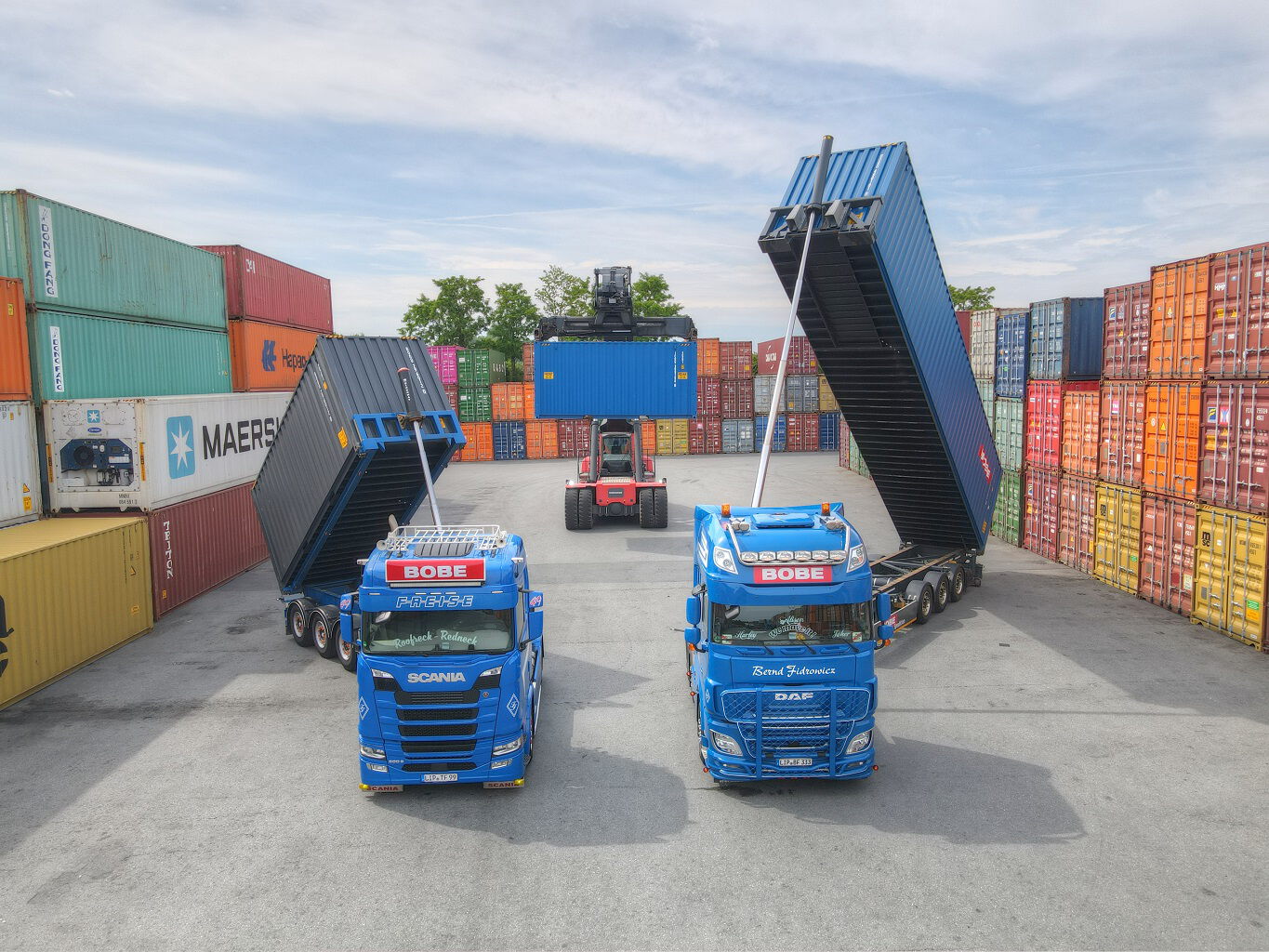 Zwei blaue BOBE-LKW mit aufgestellten Kipp-Chassis und ein Reachstacker beim Containerumschlag vor einer Kulisse aus Seecontainern – BOBE