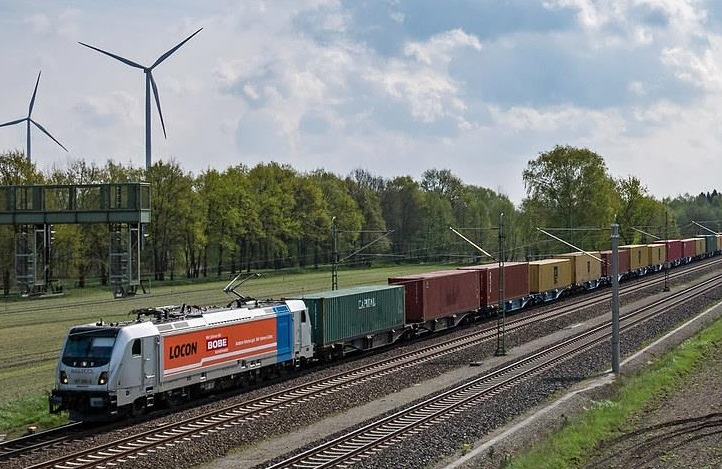 Ein langer Containerzug mit einer LOCON-Lokomotive im BOBE-Design fährt durch eine ländliche Gegend mit Windrädern – BOBE