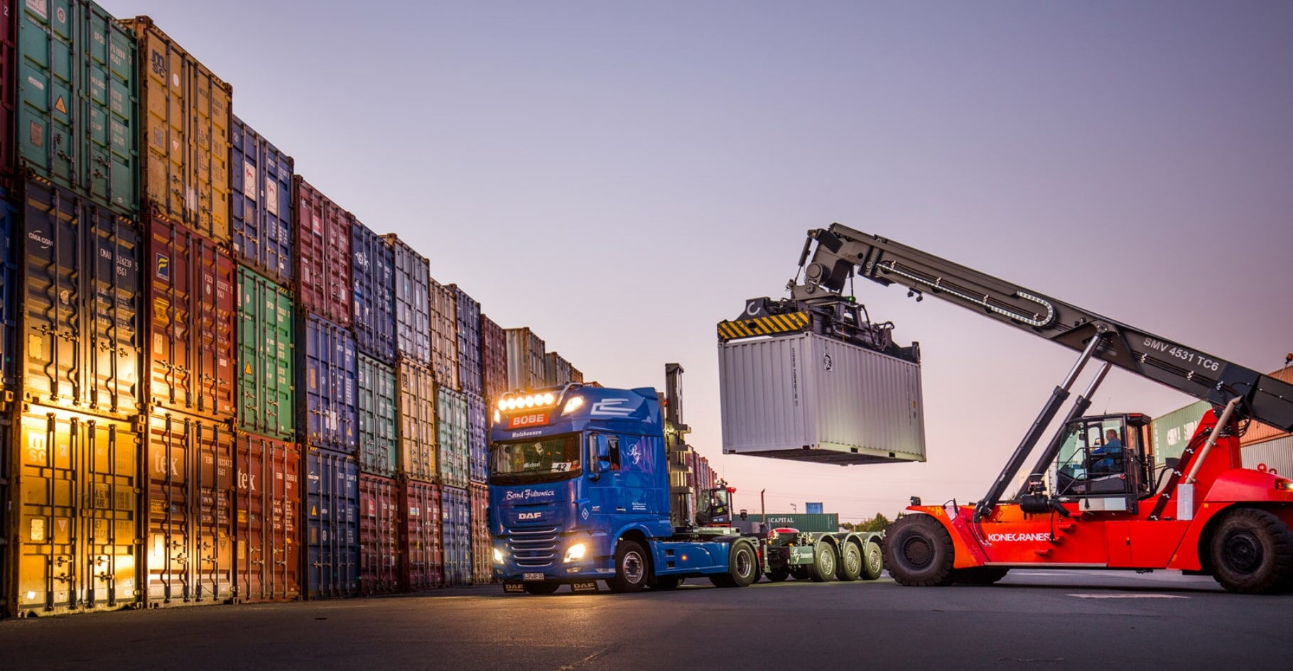 Ein Reachstacker belädt einen blauen LKW mit einem Container vor hohen Stapelreihen – BOBE