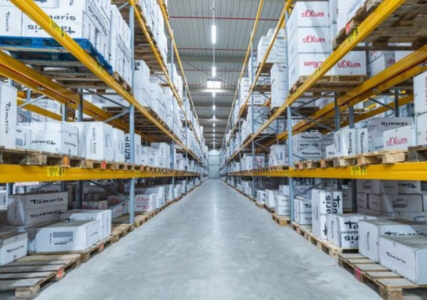 Ein langer Regalgang mit Paletten voller weißer Warenkartons namhafter Hersteller in einer hellen Logistikhalle – BOBE