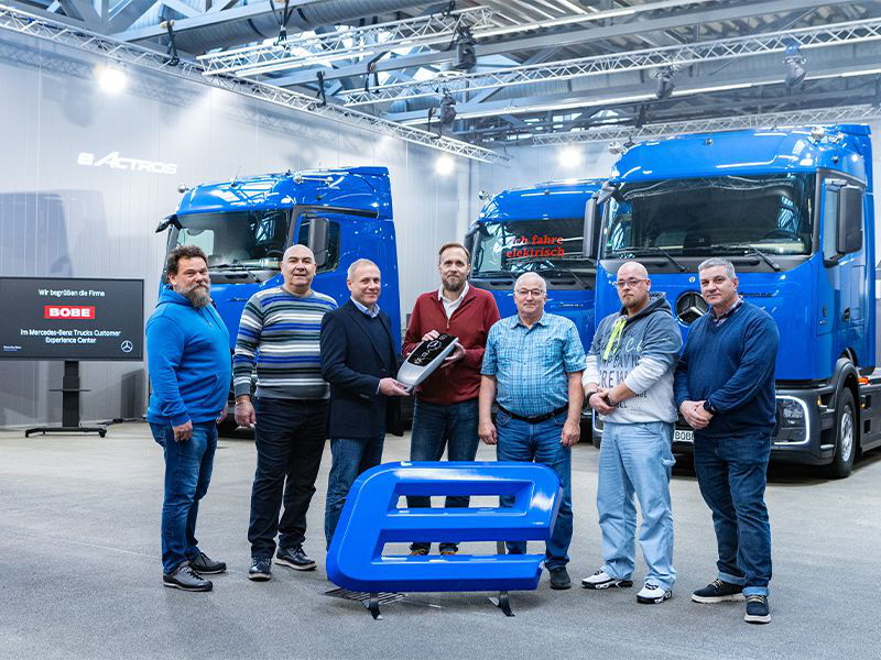 Ein Team steht bei der symbolischen Schlüsselübergabe vor blauen E-LKW in einer Auslieferungshalle – BOBE