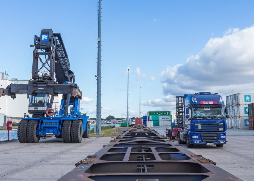Ein blauer BOBE-LKW neben einem Reachstacker bei der Be- und Entladung von Containerchassis an einem Wasserweg – BOBE