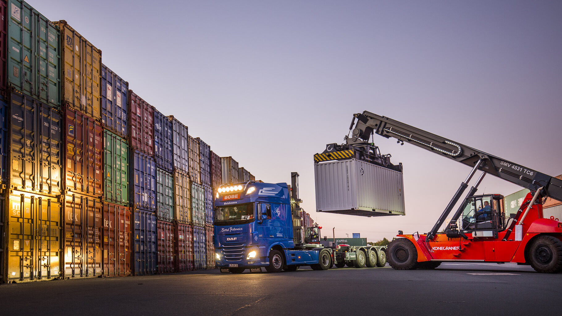 Ein blauer BOBE LKW bei der Beladung durch einen Reachstacker im Containerdepot zur Dämmerung